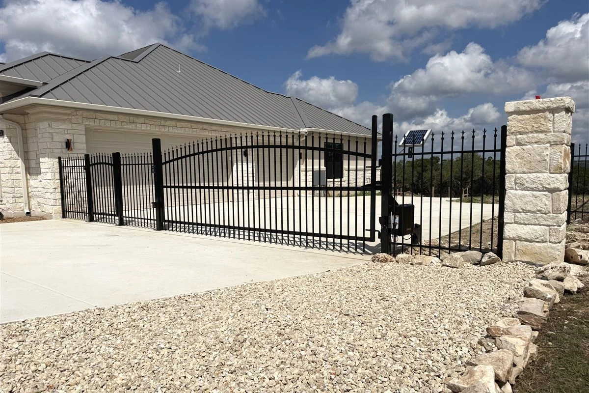 Black iron automatic driveway gate with a solar-powered gate opener and stone pillar in front of a new limestone home